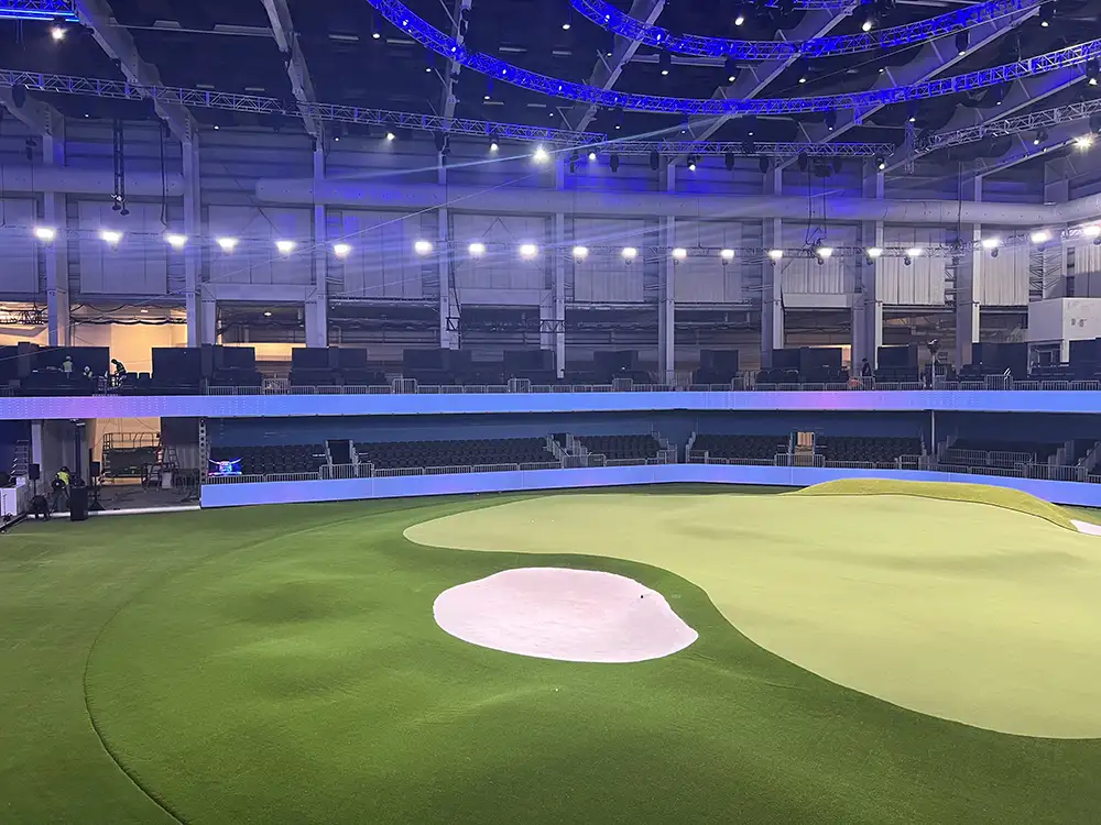 Empty indoor golf arena with a synthetic turf green, a pink sand bunker, and blue stage lighting